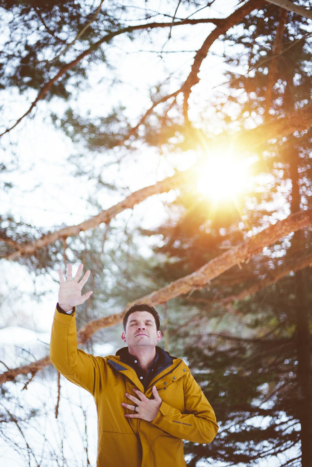 man raising left hand, right hand on chest closed eyes standing outdoor under shade of tree during daytime