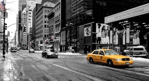 A yellow taxi drives along a city street lined with tall buildings on a gray day. The ground is lightly covered with snow, and the road appears wet. Large advertisements are visible on the building walls, and several other vehicles are present, including a bus and a black car. American flags are displayed on some of the buildings.