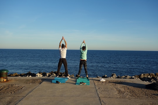 Two people are performing yoga on mats placed on a concrete surface near the ocean. They stand side by side facing the sea, both stretching their arms upwards in a symmetrical pose. There is a clear blue sky and the ocean extends to the horizon. Nearby, there are rocky edges and a single shoe placed beside one of the mats.
