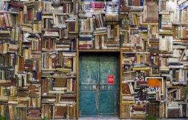 An old, weathered wall made entirely of stacked vintage books with varying colors and sizes surrounds a green, rustic wooden door. The books are tightly packed, creating a textured surface with visible spines and pages. A small red sign is affixed to the door, adding a pop of color to the muted tones.