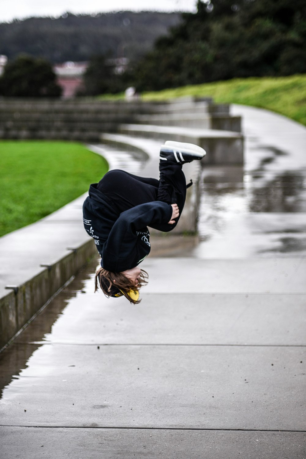 person wears black jacket and black pants doing flip on gray concrete walkway with water during daytime selective focus photography