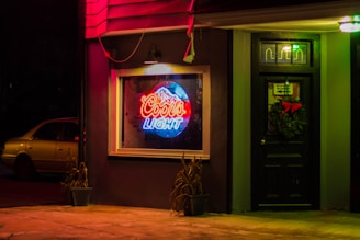 Neon Coors Light sign displayed in a window of a dimly lit building exterior at night. A black door with a wreath is next to the window, and a potted plant is placed on either side of the window. A parked car is partially visible in the background.