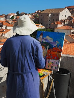 A person wearing a broad hat and a blue coat is painting a colorful cityscape on a canvas, with red-tiled rooftops and stone buildings in the background. The scene reflects a vibrant blend of colors, capturing the essence of a sunny day.