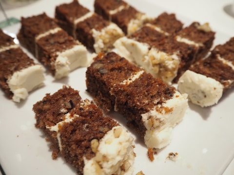 Sliced cake pieces with a dark, dense appearance are arranged on a white plate. Each piece is layered with a creamy filling, and some slices have visible nut pieces or seeds. The texture appears rich, with a blend of moist cake and smooth cream.