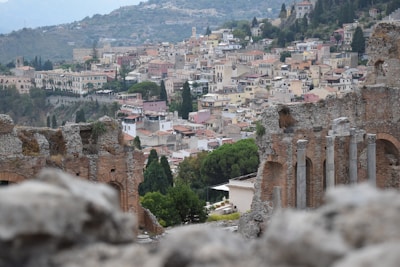 A scenic view features the ancient ruins of a theater with weathered stone columns and arches in the foreground. The background reveals a colorful hillside town, with numerous closely packed buildings and varied rooflines nestled among lush greenery.
