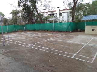 A concrete badminton court is outlined with white lines. It is located outdoors, surrounded by trees and a building in the background. A green mesh fence encloses the area. Metal poles for the net are seen on the court. The surface is slightly worn, with some scattered leaves and debris.