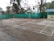A concrete badminton court is outlined with white lines. It is located outdoors, surrounded by trees and a building in the background. A green mesh fence encloses the area. Metal poles for the net are seen on the court. The surface is slightly worn, with some scattered leaves and debris.