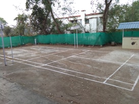 A concrete badminton court is outlined with white lines. It is located outdoors, surrounded by trees and a building in the background. A green mesh fence encloses the area. Metal poles for the net are seen on the court. The surface is slightly worn, with some scattered leaves and debris.
