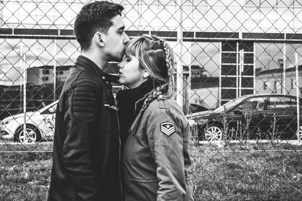grayscale photo of man kissing forehead of woman near fence