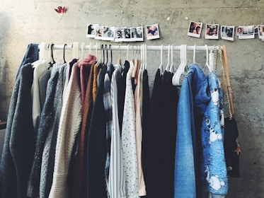 A collection of various clothing items is neatly arranged on hangers along a horizontal rack against a textured gray wall. Above the rack, a string of small polaroid photos is pinned, adding a personal touch to the space. The clothing includes a range of sweaters, shirts, and jackets in various colors from neutrals to blues.