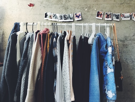A collection of various clothing items is neatly arranged on hangers along a horizontal rack against a textured gray wall. Above the rack, a string of small polaroid photos is pinned, adding a personal touch to the space. The clothing includes a range of sweaters, shirts, and jackets in various colors from neutrals to blues.