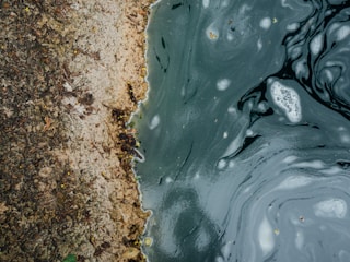 A textured area of soil or sand meets the swirling, multicolored surface of water, possibly polluted, with visible bubbles and debris. The scene has an abstract, natural feel with earthy tones and a contrast between solid and fluid forms.
