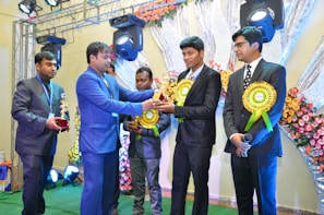 A group of men in formal suits stand on a stage decorated with flowers, where one man presents a trophy to another. Bright lighting and equipment are visible overhead, creating a festive atmosphere.