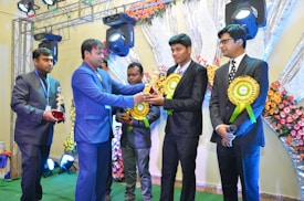 A group of men in formal suits stand on a stage decorated with flowers, where one man presents a trophy to another. Bright lighting and equipment are visible overhead, creating a festive atmosphere.