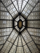 An intricately designed ceiling featuring a geometric pattern of intersecting lines and embossed metallic panels. The ceiling is divided into sections by dark beams, creating a symmetrical appearance that draws the eye towards a central, elongated hexagonal opening. The panels have an ornate, antiquated look with circular motifs, and they reflect a soft, warm light.