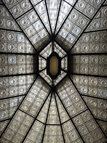 An intricately designed ceiling featuring a geometric pattern of intersecting lines and embossed metallic panels. The ceiling is divided into sections by dark beams, creating a symmetrical appearance that draws the eye towards a central, elongated hexagonal opening. The panels have an ornate, antiquated look with circular motifs, and they reflect a soft, warm light.