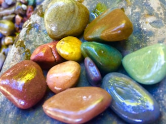 A collection of colorful, smooth pebbles arranged on a larger rock surface. The pebbles vary in shades of green, yellow, red, and brown, reflecting light with a glossy finish.