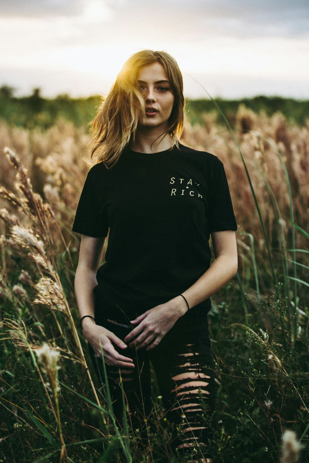 昼間、茶色の小麦畑に立つ黒のクルーネックTシャツとユーズド黒のボトムのセレクティブフォーカス写真
