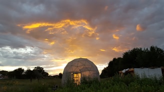 A geodesic dome structure is set against a dramatic sunset sky, with vibrant golden and orange hues illuminating the clouds. The dome is situated in a lush green landscape with trees and a small building in the background.