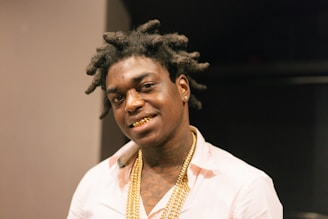 A person with styled hair wearing a light-colored shirt and gold chains, smiling with a visible gold grill on their teeth. The background is dimly lit, creating a focus on the subject.