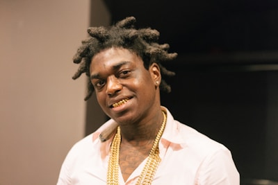 A person with styled hair wearing a light-colored shirt and gold chains, smiling with a visible gold grill on their teeth. The background is dimly lit, creating a focus on the subject.