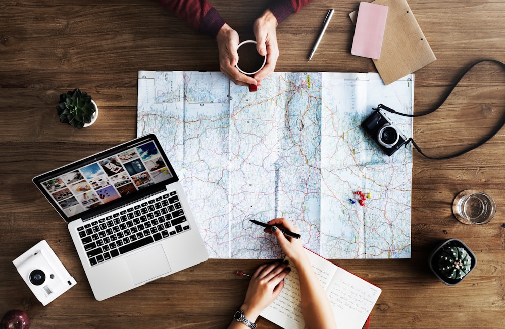 An overhead shot of two people planning a trip with a map and a laptop on a wooden surface