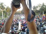 Crowd gathered at a lively outdoor event with banners and cameras.