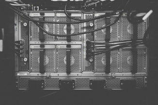 A monochrome photograph of a server rack with multiple units. The image shows intricate patterns of ventilation grids and various cables connected to the hardware. Small indicator lights suggest the equipment is operational.
