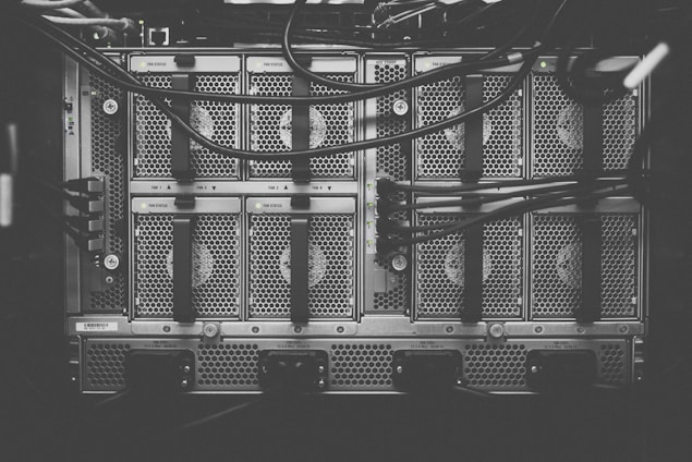 A monochrome photograph of a server rack with multiple units. The image shows intricate patterns of ventilation grids and various cables connected to the hardware. Small indicator lights suggest the equipment is operational.