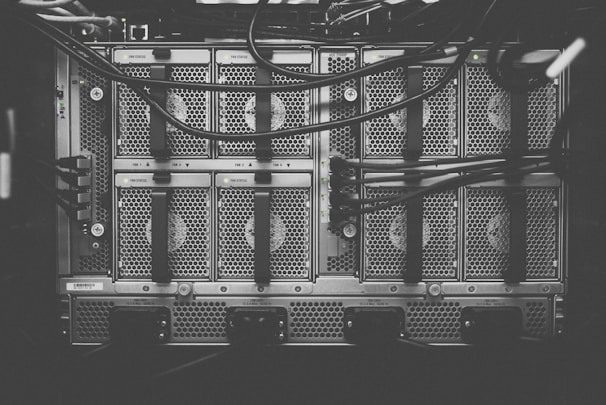 A monochrome photograph of a server rack with multiple units. The image shows intricate patterns of ventilation grids and various cables connected to the hardware. Small indicator lights suggest the equipment is operational.