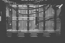 A monochrome photograph of a server rack with multiple units. The image shows intricate patterns of ventilation grids and various cables connected to the hardware. Small indicator lights suggest the equipment is operational.