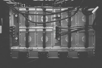 A monochrome photograph of a server rack with multiple units. The image shows intricate patterns of ventilation grids and various cables connected to the hardware. Small indicator lights suggest the equipment is operational.