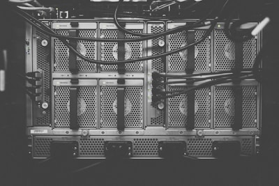 A monochrome photograph of a server rack with multiple units. The image shows intricate patterns of ventilation grids and various cables connected to the hardware. Small indicator lights suggest the equipment is operational.
