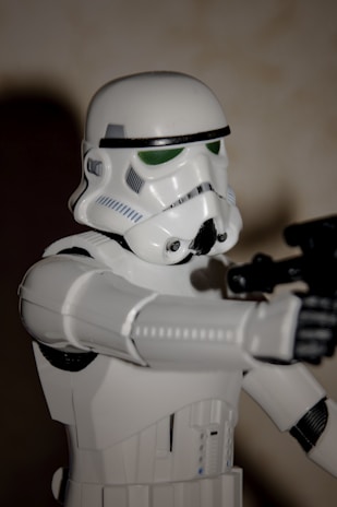A detailed action figure of a stormtrooper from the Star Wars franchise is posed with a blaster in its hand. The figure is primarily white with black and grey accents, standing at an angle against a plain background.