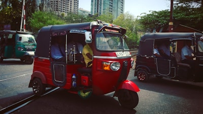 A vibrant red auto rickshaw is stationed on a street near tram tracks. The vehicle is occupied by several passengers, and it stands in the foreground of an urban setting. Buildings and lush greenery can be seen in the background, suggesting a bustling city environment. Other tuk-tuks are visible on the road, contributing to a busy and lively atmosphere.