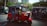 A vibrant red auto rickshaw is stationed on a street near tram tracks. The vehicle is occupied by several passengers, and it stands in the foreground of an urban setting. Buildings and lush greenery can be seen in the background, suggesting a bustling city environment. Other tuk-tuks are visible on the road, contributing to a busy and lively atmosphere.