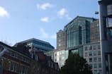 A modern cityscape featuring several buildings, including a large structure with blue-tinted glass windows and an arched design. The sky is clear with a few clouds, and there is a sculpture of a white animal visible on the left.