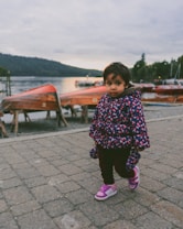 A young child dressed in a colorful jacket with floral patterns and pink shoes is standing on a paved walkway. In the background, there are several wooden rowboats near a calm body of water, surrounded by forested hills under a cloudy sky.