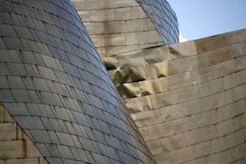 Curved and angular metallic surfaces form an abstract architectural structure, with panels reflecting light and creating a dynamic play of shadows. The structure features overlapping sections and an intricate design with a sleek and modern aesthetic.