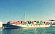 A large cargo ship is sailing across the water, laden with numerous colorful shipping containers stacked in rows. The skyline is visible in the background, along with a suspension bridge stretching across the scene. The ship bears the name Cosco Ningbo and the water reflects its image under a clear blue sky.