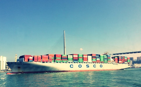 A large cargo ship is sailing across the water, laden with numerous colorful shipping containers stacked in rows. The skyline is visible in the background, along with a suspension bridge stretching across the scene. The ship bears the name Cosco Ningbo and the water reflects its image under a clear blue sky.