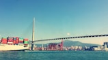 A large cargo ship laden with colorful shipping containers sails beneath a modern cable-stayed bridge. In the background, a bustling port with cranes and a skyline of urban buildings set against a mountainous landscape. The sky is clear, contributing to the vivid colors in the scene.