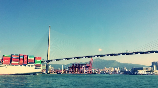A large cargo ship laden with colorful shipping containers sails beneath a modern cable-stayed bridge. In the background, a bustling port with cranes and a skyline of urban buildings set against a mountainous landscape. The sky is clear, contributing to the vivid colors in the scene.