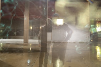 A blurred reflection of a person is seen against a glass pane with visible streaks and light patterns. There is a brick wall in the background, and the ground appears wet as if after rain. The scene has an abstract and dreamy quality due to the lighting and reflections.
