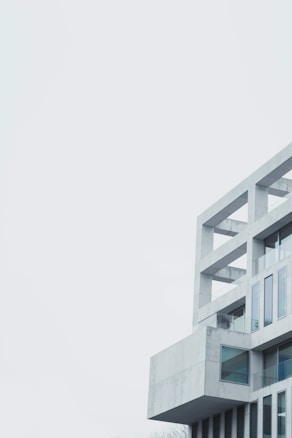 A modern, minimalist building with geometric lines and multiple rectangular glass windows, constructed from light gray concrete. The structure features a series of open, square frames near the top, creating a sense of openness and space.