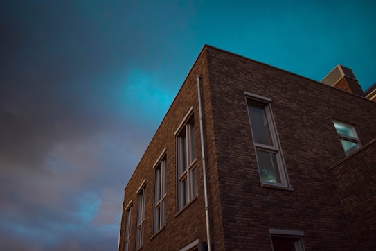 The image features a brick building viewed from a low angle, highlighting the corner where two walls meet. The sky above is dramatic, filled with dark clouds transitioning to a deep teal hue. The windows on the building reflect some light, adding to the contrast between the structure and the sky.