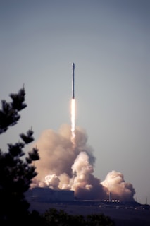 A rocket is launching into the sky, leaving behind a trail of smoke and fire as it ascends. The base of the launch pad is surrounded by clouds of smoke, indicating a recent lift-off. A tree is partially visible in the foreground, framing the scene.