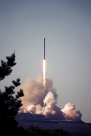A rocket is launching into the sky, leaving behind a trail of smoke and fire as it ascends. The base of the launch pad is surrounded by clouds of smoke, indicating a recent lift-off. A tree is partially visible in the foreground, framing the scene.