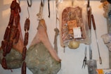 Various types of cured meats, including sausages, hams, and a packaged pancetta, are hanging from hooks. The meats are arranged on a wall, showing a rustic and artisanal display. Cooking tools with wooden handles are also hanging next to the meats.