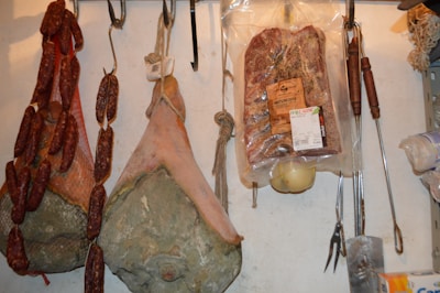 Various types of cured meats, including sausages, hams, and a packaged pancetta, are hanging from hooks. The meats are arranged on a wall, showing a rustic and artisanal display. Cooking tools with wooden handles are also hanging next to the meats.